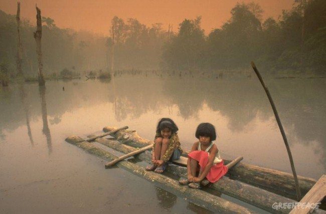 Two small children sit on a raft on a small lake amid smoke and haze while a forest fire rages in the background